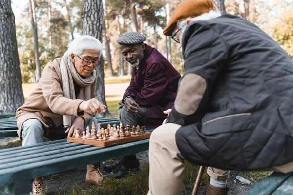 Старший азиат играет в шахматы с межрасовыми друзьями в осеннем парке — стоковое фото