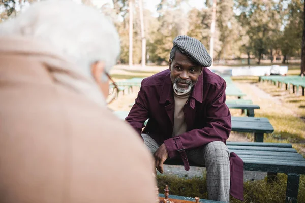 Hombre afroamericano mayor mirando a amigo borroso cerca de ajedrez en el parque - foto de stock