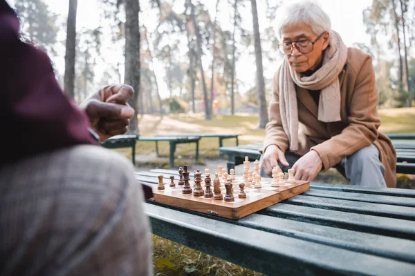Senior asian man sitting near chess and blurred african american friend in autumn park — Stock Photo
