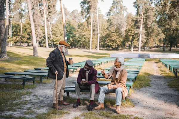 Multiethnic men with smartphone and coffee to go talking in autumn park — Stock Photo