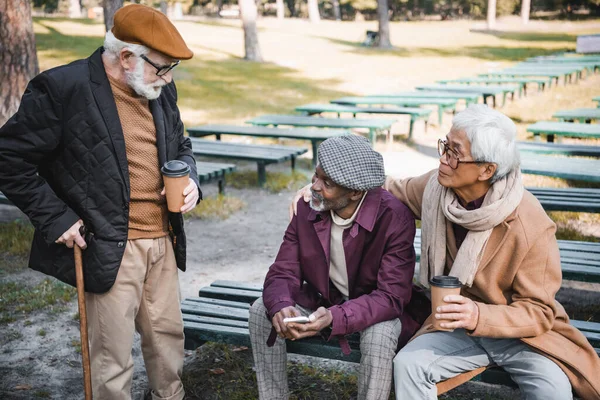 Hombres mayores multiculturales con café y teléfono inteligente pasar tiempo en el parque - foto de stock