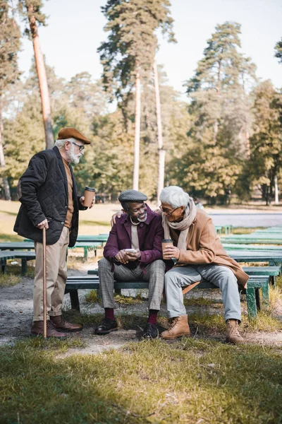 Веселі багатоетнічні чоловіки з паперовою чашкою та смартфоном сидять на лавці біля друга в парку — стокове фото
