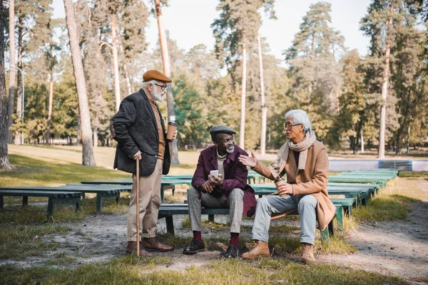 Senior asian man with coffee to go pointing with hand while talking to interracial friends with smartphone in park — Stock Photo