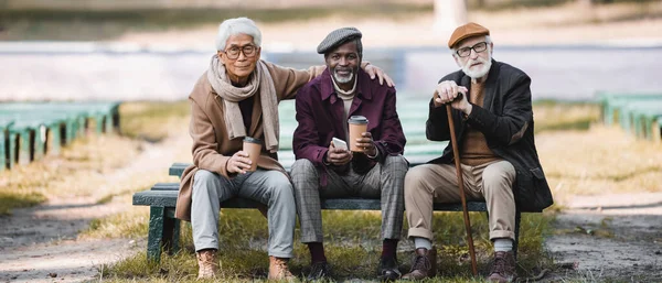 Uomini anziani multietnici con bicchieri di carta, smartphone e bastone da passeggio seduti su panchina nel parco, banner — Foto stock