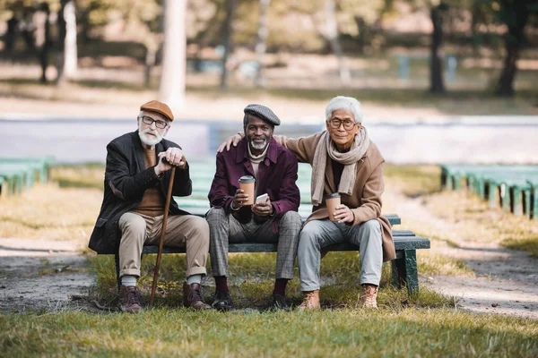 Interracial elderly friends with coffee to go and smartphone sitting on bench in park — Stock Photo