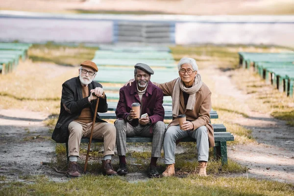 Multiethnische Senioren mit Pappbechern und Smartphone sitzen auf Bank neben Freund mit Gehstock — Stockfoto