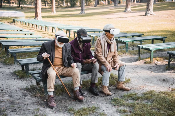 Elderly multiethnic friends with smartphone and vr headsets sitting on bench in park — Stock Photo