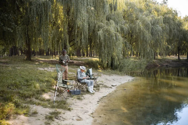 Africano americano uomo holding occhiali e termo coppa vicino asiatico amico in abito da pesca vicino lago — Foto stock