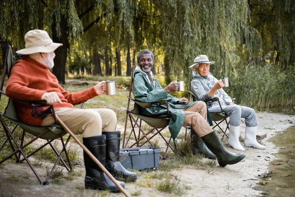 Sonrientes hombres mayores multiculturales en traje de pesca con copas termo sentado cerca del lago en el parque - foto de stock