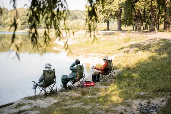 Interracial men in fishing outfit sitting on chairs near lake — Stock Photo