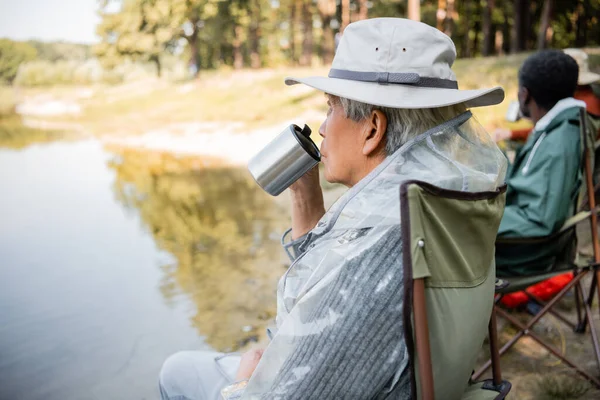 Senior asiático hombre celebración thermo taza cerca borrosa amigos en traje de pesca cerca de lago - foto de stock