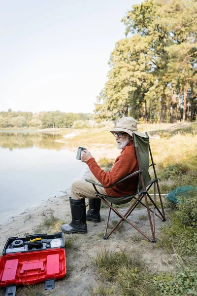 Homem sênior em roupa de pesca segurando copo termo perto de caixa de ferramentas e lago — Fotografia de Stock