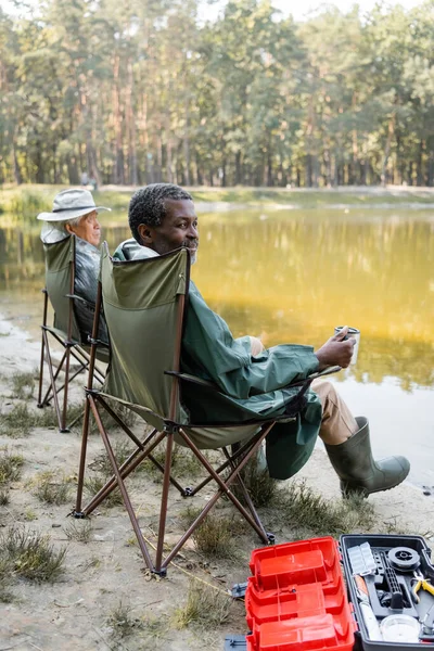 African american man holding thermo cup near asian friend in fishing outfit near toolbox and lake — Stock Photo