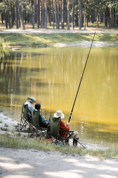 Amigos mayores multiétnicos con caña de pescar sentados cerca del lago en el parque - foto de stock