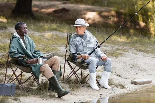 Asian man with fishing rod looking at african american friend with thermo cup near lake — Stock Photo