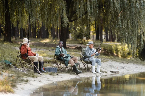 Multiethnic senior men with fishing rods holding thermo cups near lake — Stock Photo