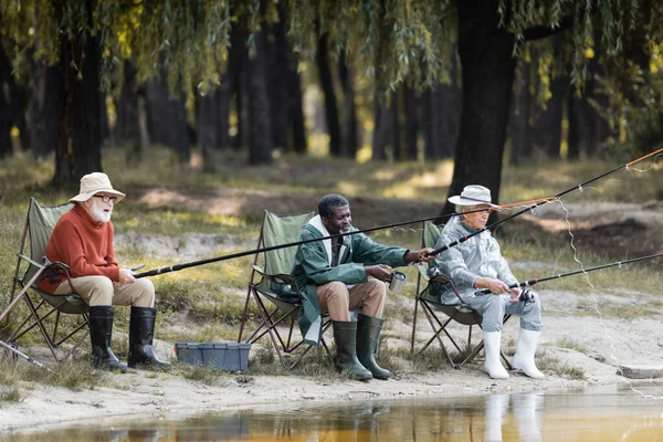 Multiethnic elderly men with fishing rods spending time near lake — Stock Photo