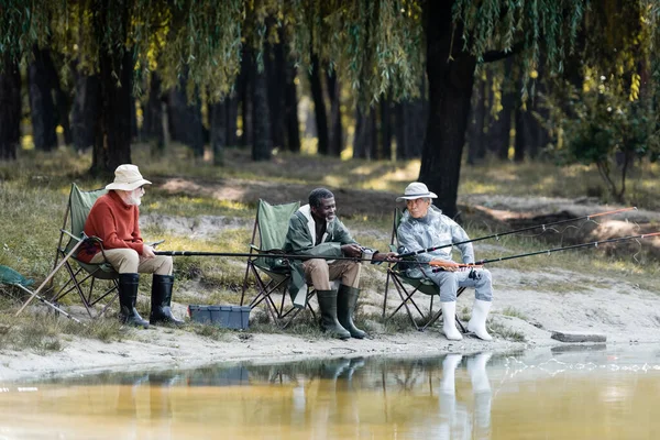 Sorridente uomo africano americano in stivali di gomma pesca vicino amici interrazziali e cassetta degli attrezzi — Foto stock