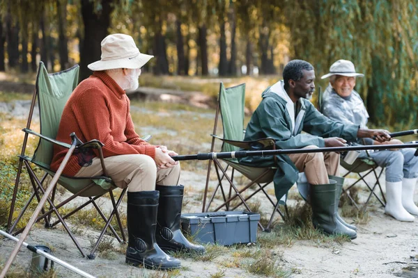 Älterer Mann mit Angelrute sitzt in der Nähe von Werkzeugkiste und Freunden im Freien — Stockfoto