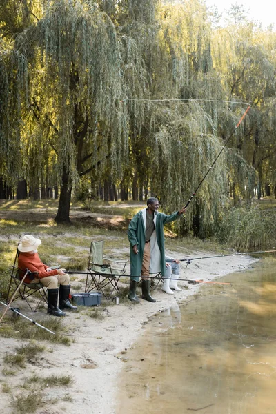 Hombre afroamericano pescando cerca de amigos mayores y caja de herramientas al aire libre - foto de stock