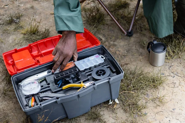 Vista recortada del hombre afroamericano tomando herramienta de la caja de herramientas durante la pesca al aire libre - foto de stock