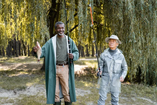 Smiling african american man holding fish near asian friend with fishing rod outdoors — Stock Photo
