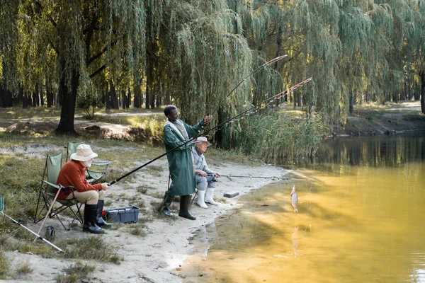 Hombre afroamericano captura de peces cerca de amigos interracial con cañas de pescar y lago - foto de stock