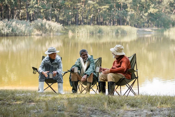 Позитивные межрасовые мужчины в рыболовной экипировке держат термостаты на стульях на берегу озера — стоковое фото