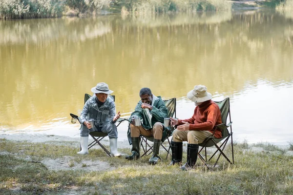 Positive interracial senior men in fishing outfit holding thermo cups while spending time outdoors — Stock Photo