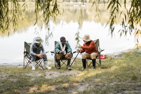 Senior man with thermo cup talking to interracial friends in fishing outfit on lake coast — Stock Photo