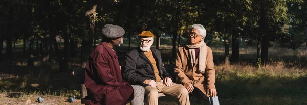 Ancianos hombres multiétnicos hablando en el banco en el parque, pancarta - foto de stock