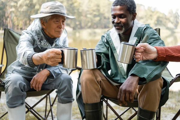 Blurred senior friends in fishing outfit holding thermo cups outdoors — Stock Photo