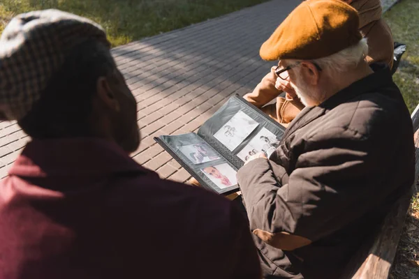 Elderly man holding photo album near blurred interracial friends on bench in park — Stock Photo