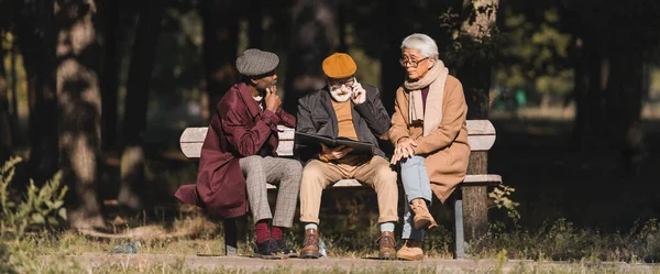 Hombre mayor con álbum de fotos hablando en el teléfono inteligente cerca de amigos interracial en el parque, pancarta - foto de stock