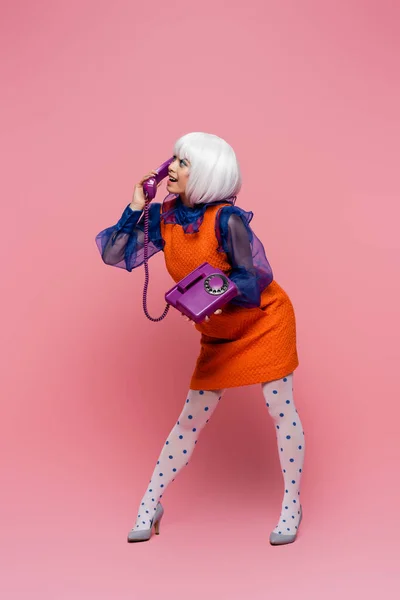 Smiling asian woman in vintage dress and white wig talking on telephone on pink background — Stock Photo