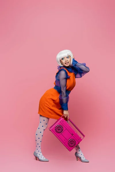 Mujer asiática alegre en peluca blanca y vestido retro sosteniendo boombox sobre fondo rosa - foto de stock