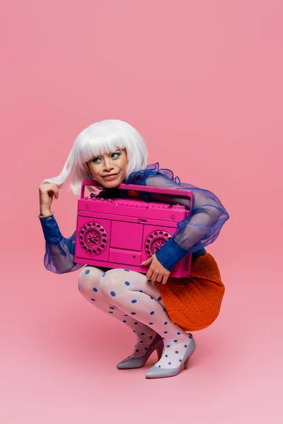 Sonriendo mujer asiática tocando peluca blanca y sosteniendo boombox vintage sobre fondo rosa - foto de stock