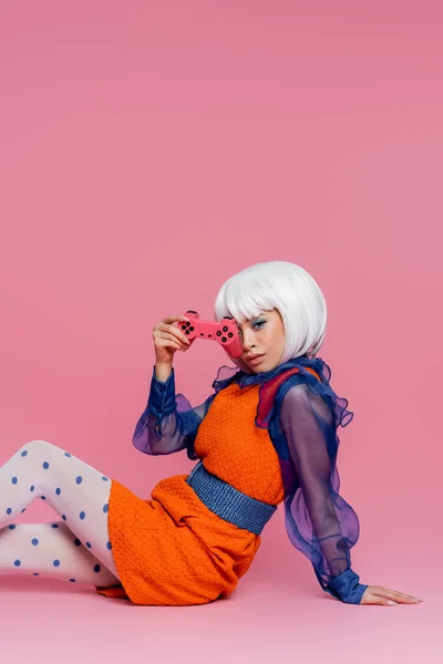 KYIV, UCRANIA - 10 de diciembre de 2020: Modelo de arte pop asiático que sostiene el joystick mientras está sentado sobre un fondo rosa - foto de stock