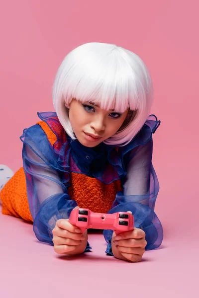 KYIV, UKRAINE - DECEMBER 10, 2020: Stylish asian woman in wig holding joystick while lying on pink background — Stock Photo