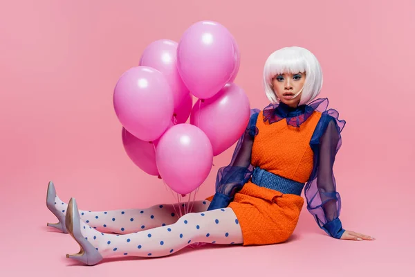 Stylish asian pop art woman looking at camera while sitting near balloons on pink background — Stock Photo