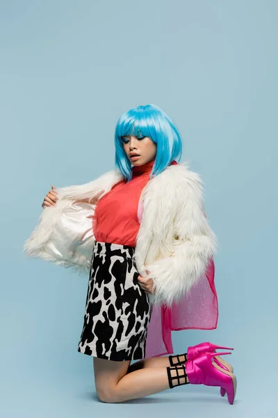 Asian woman in wig adjusting furry jacket while standing on knees on blue background — Stock Photo
