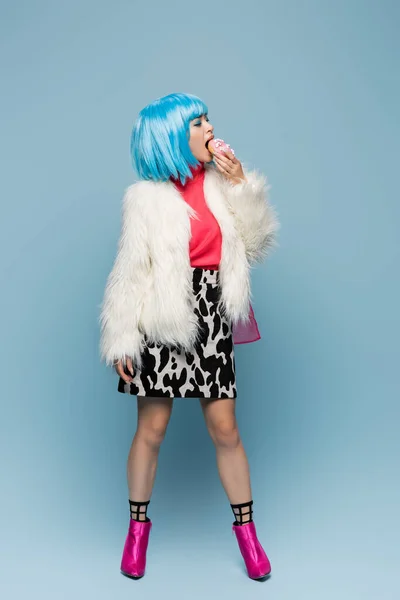 Full length of stylish asian pop art woman eating donut on blue background — Stock Photo