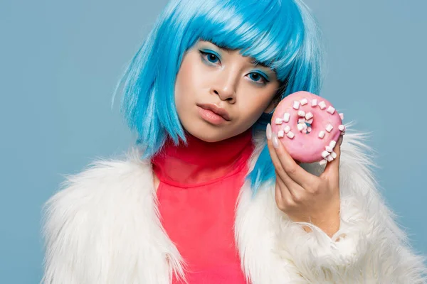 Mujer asiática en peluca y chaqueta mullida sosteniendo donut y mirando a la cámara aislada en azul - foto de stock
