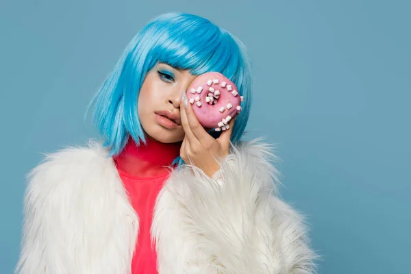 Moda joven asiático mujer en mullido chaqueta celebración donut cerca de cara aislado en azul - foto de stock