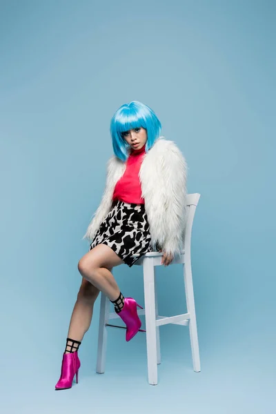 Young asian woman in fluffy jacket and heels sitting on chair on blue background — Stock Photo