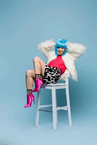 Позитивная азиатка в ярком парике и на каблуках сидит на стуле на синем фоне — стоковое фото