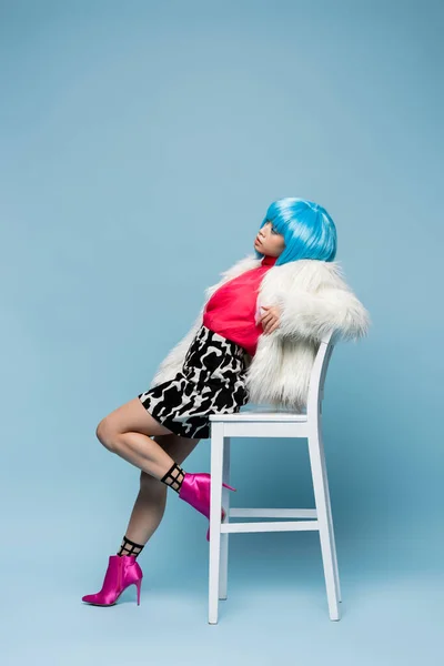 Mujer de moda en chaqueta mullida y peluca brillante posando cerca de la silla sobre fondo azul - foto de stock