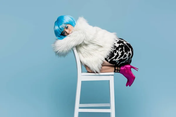 Stylish asian woman in wig, heels and fluffy jacket posing on chair isolated on blue — Stock Photo