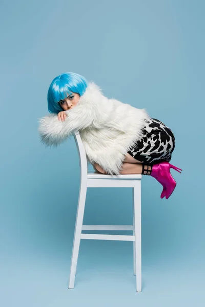 Stylish asian woman in heeled shoes and fluffy jacket posing on chair on blue background — Stock Photo