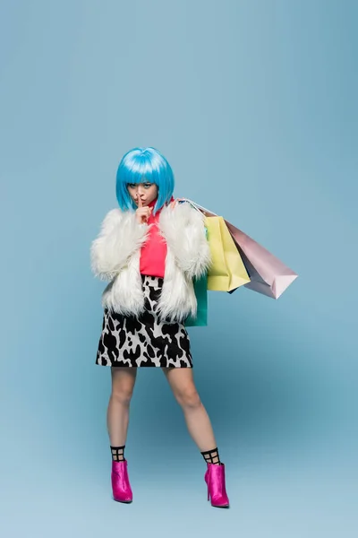 Asian woman in pop art style holding shopping bags and showing secret gesture on blue background — Stock Photo